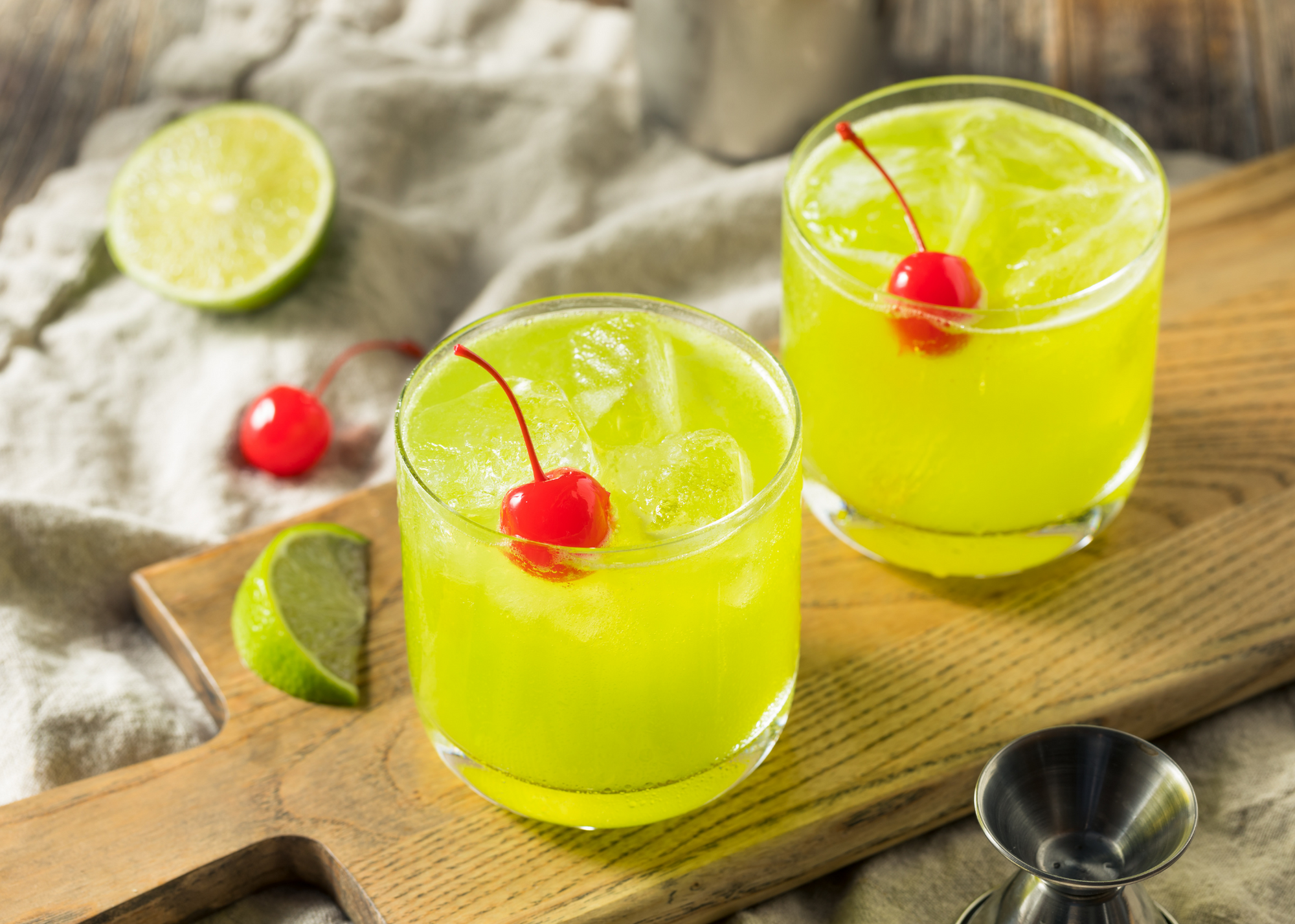 Two Midori Sours with cherry garnish on a cutting board with limes and maraschino cherries and jigger